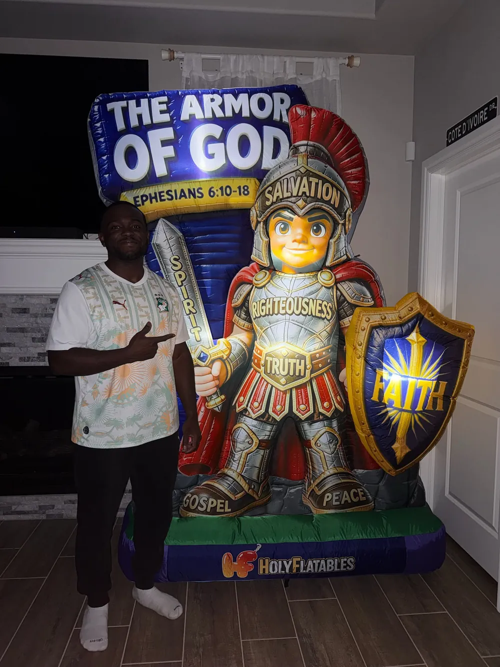 The Armor of God inflatable standing in a living room, with a smiling man in a white football-style jersey pointing at it